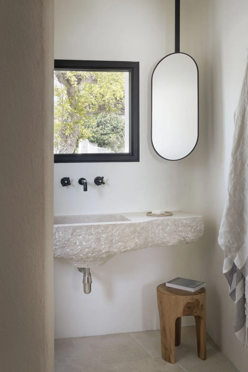 Minimalist bathroom with a stone sink, black faucets, a small wooden stool with a book, an oval mirror, and a window showing greenery outside.