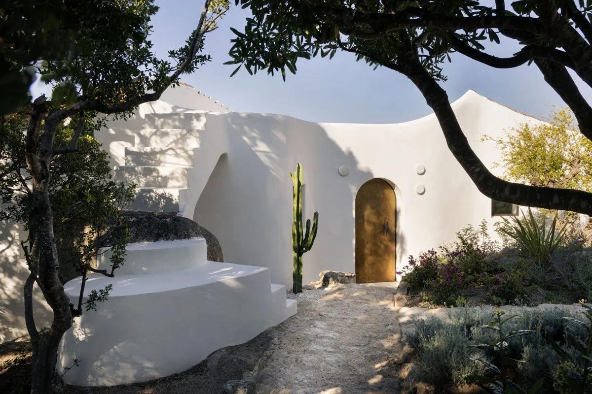 A white, Mediterranean-style building with curved walls and stairs, surrounded by green trees and plants, including a cactus near an arched doorway, with a dirt pathway leading to the door.