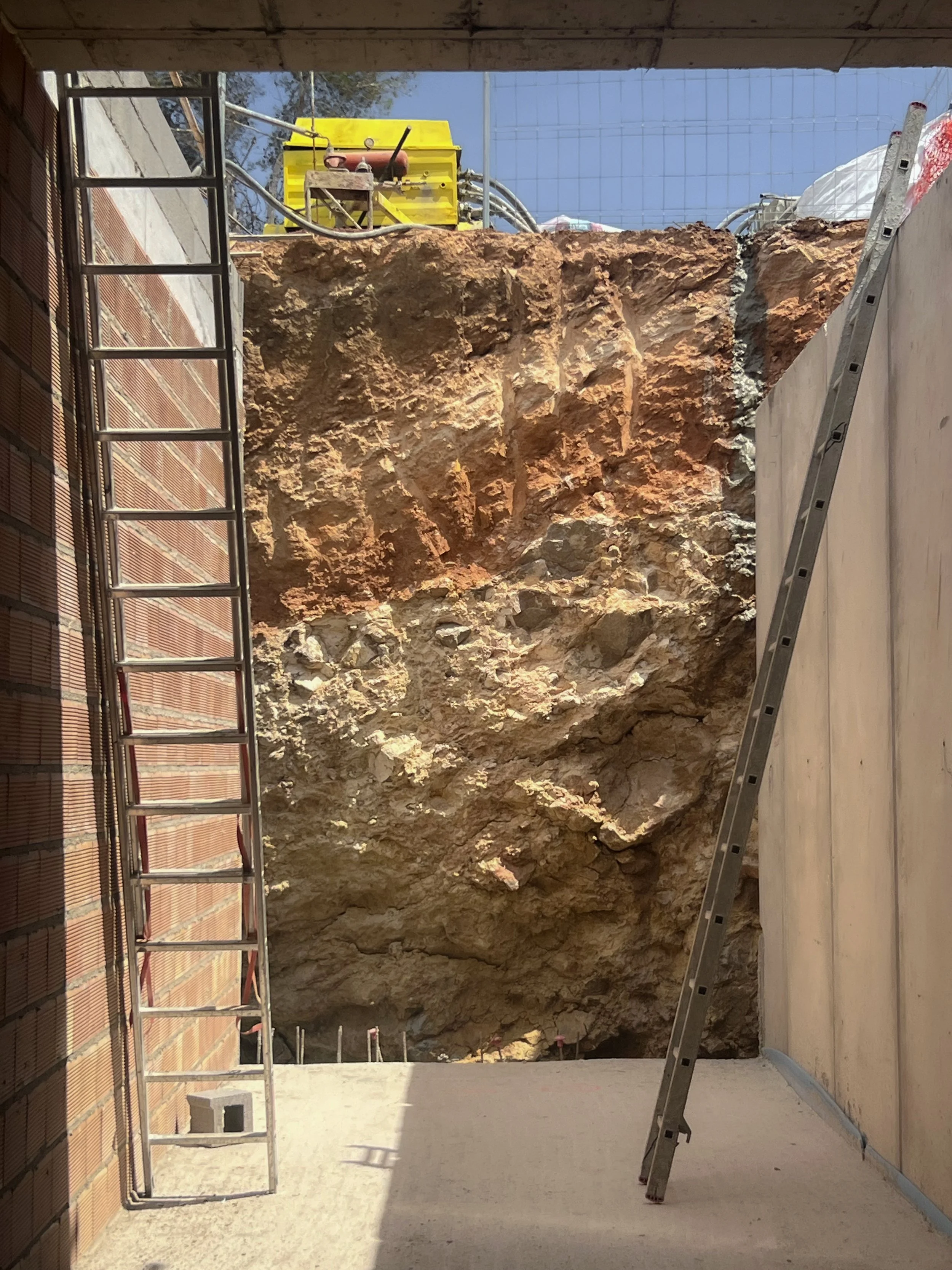 Construction site with a large excavation exposing a rock face, two ladders leaning against walls, and construction equipment at the top of the excavation.