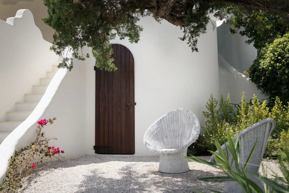 Outdoor seating area with white wicker chairs, white gravel ground, white stucco wall with dark wooden door, lush green plants, and a tree providing shade.