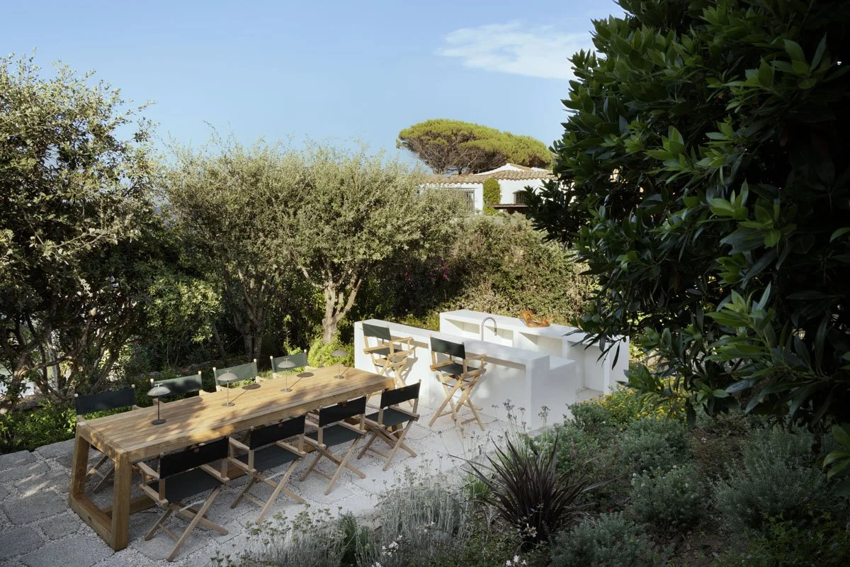 Outdoor patio with wooden dining table, black chairs, and a white outdoor kitchen with a sink, surrounded by lush green trees and plants in a sunny setting.
