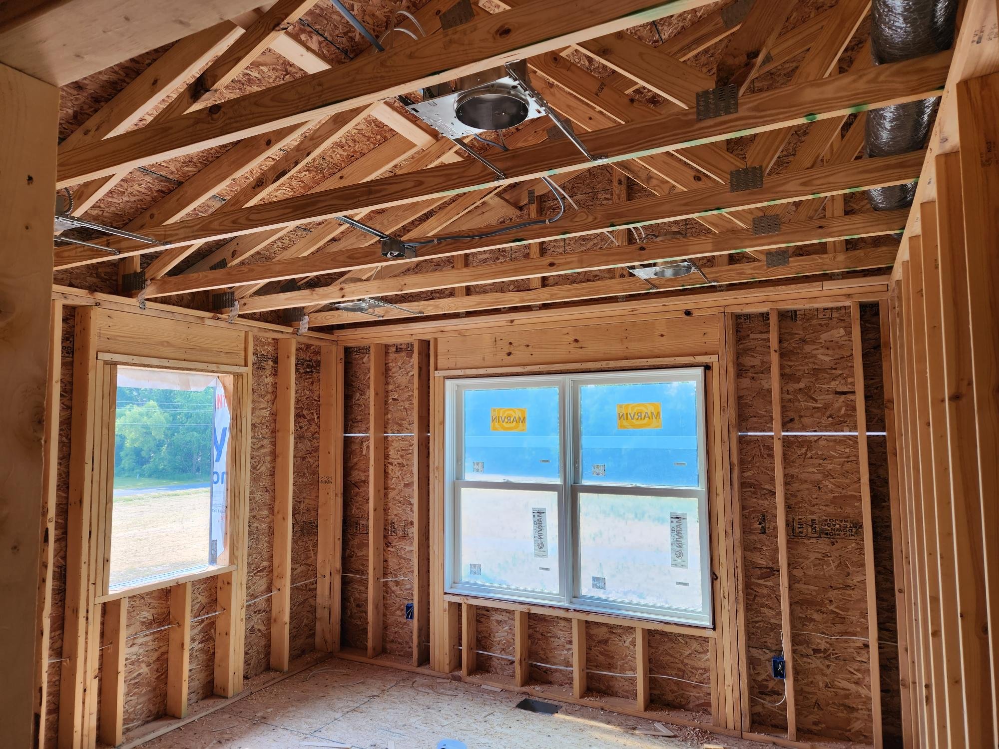 Interior of a house under construction with exposed wooden framing, roofing, and windows, showing unfinished walls and roof structure.