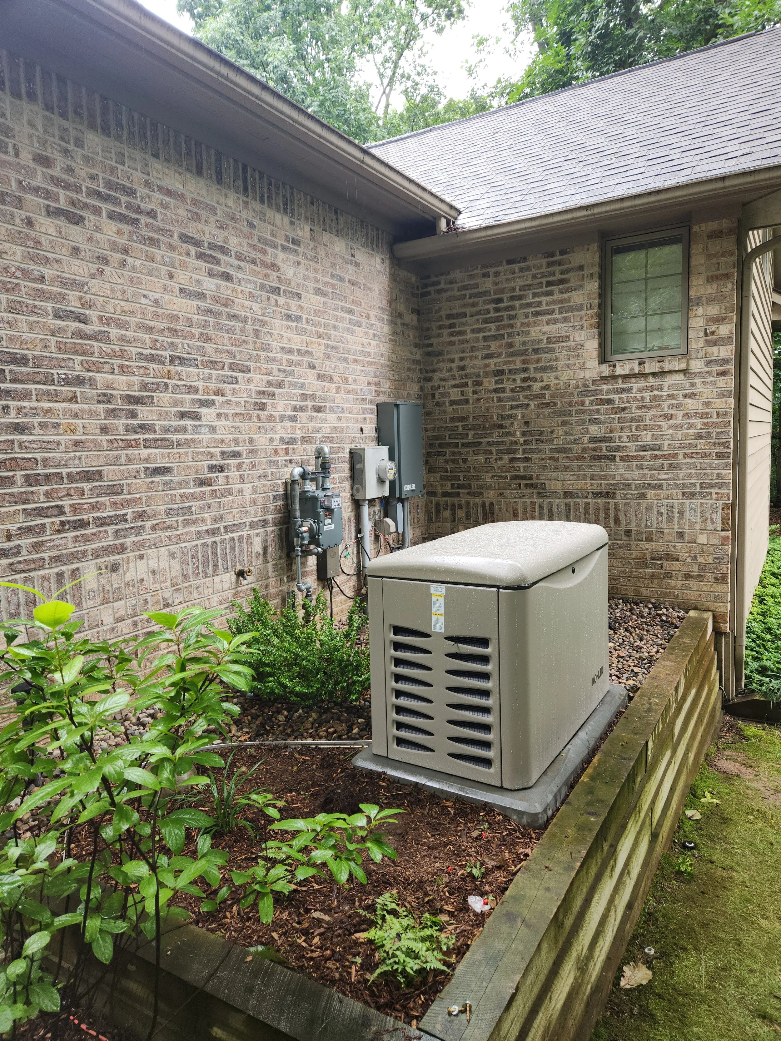 Outdoor area next to a brick house with a small window, a gray utility box, and a beige appliance in a wooden raised garden bed containing green plants and soil, surrounded by trees.