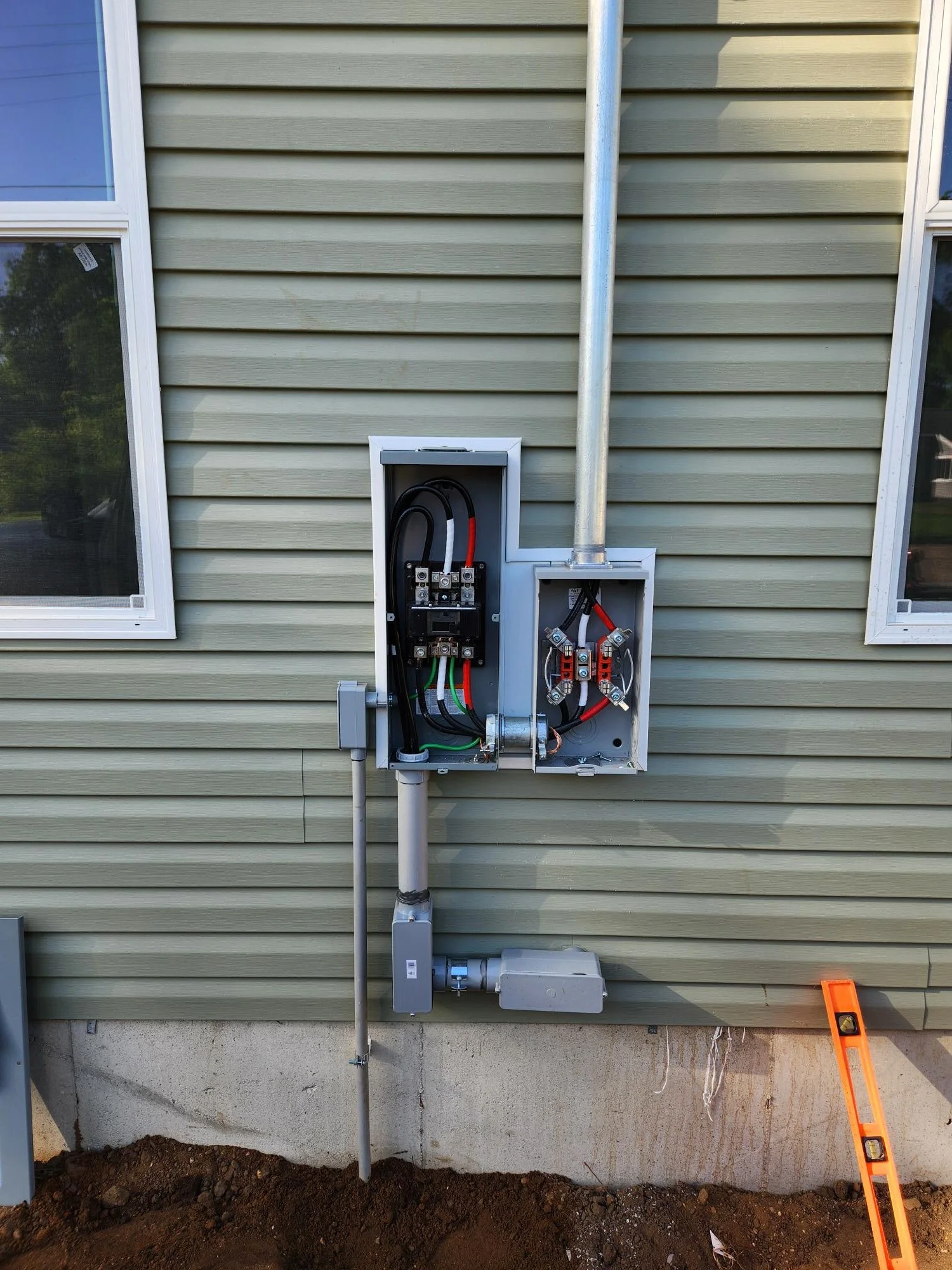 An outdoor view of an electrical meter box and conduit box mounted on a house with beige vinyl siding. The electrical panel is open, showing wiring inside. A level tool rests on the ground on the right, and a metal pipe runs vertically along the wall.