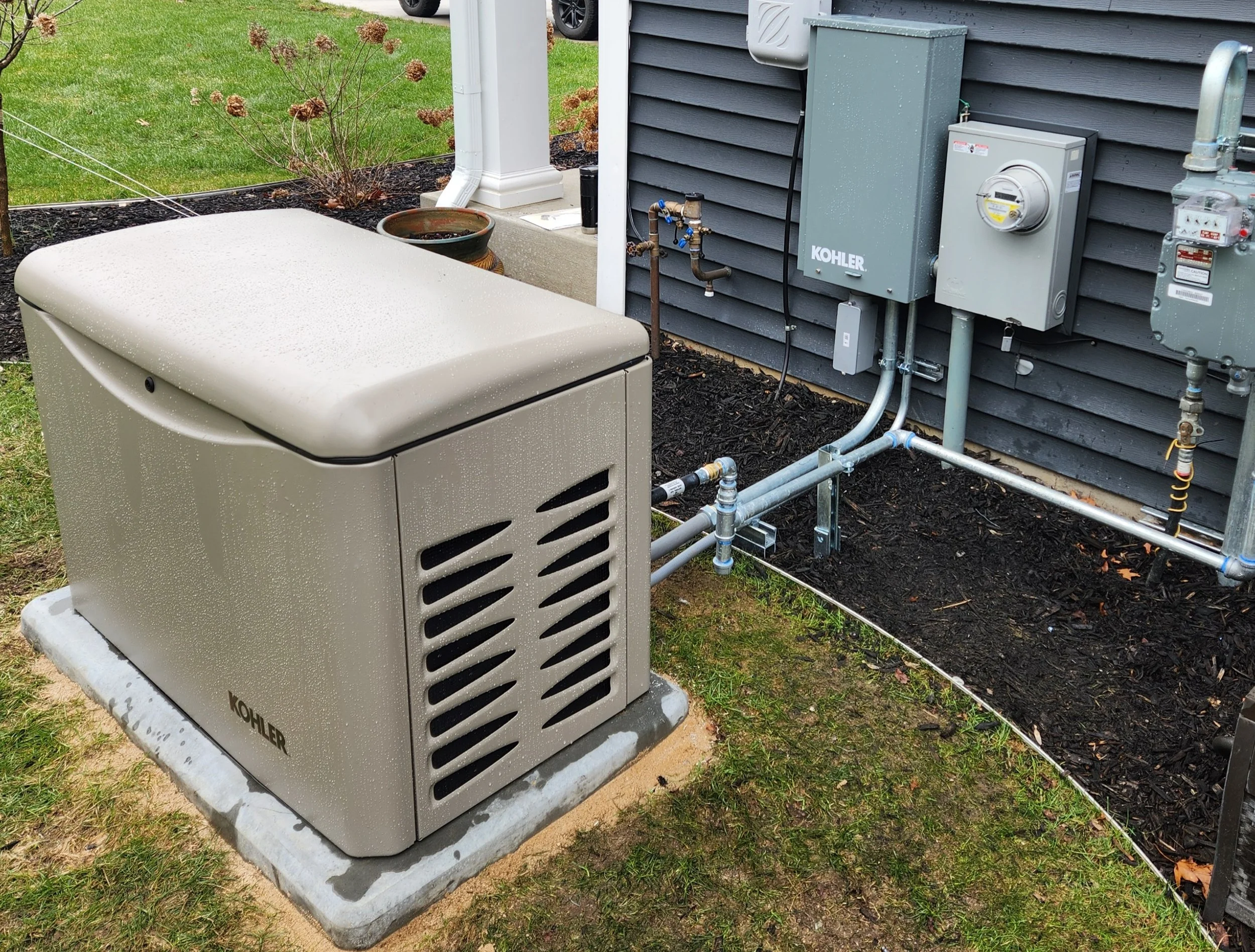Outdoor view of HVAC system unit next to house with electrical boxes and pipes on siding, on a concrete slab with grass and plants nearby.