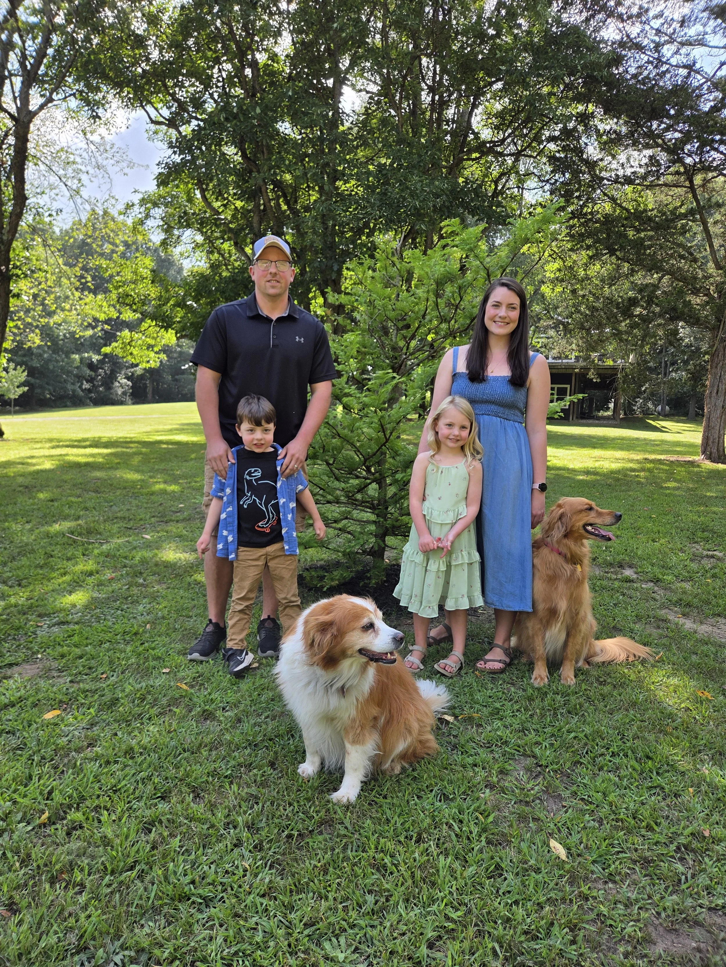 A family photo of a man, woman, and two children with two dogs in a park. They are standing on grass with trees in the background on a sunny day.
