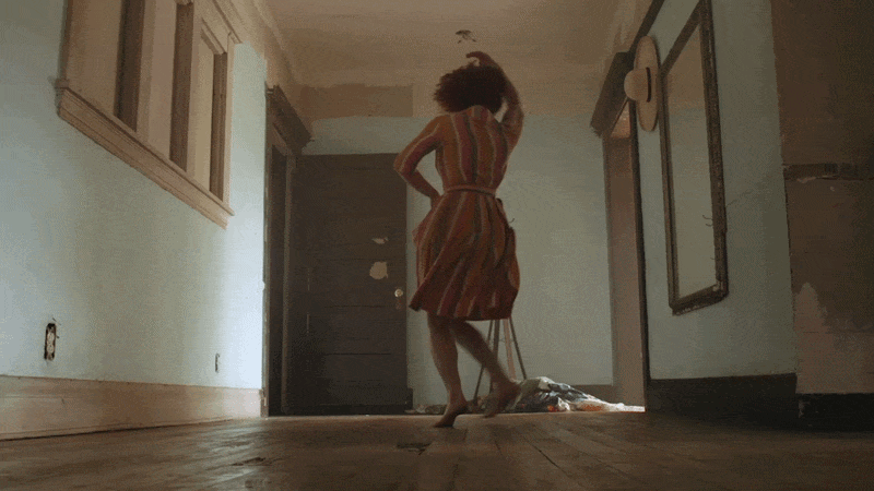 A woman in a striped dress dancing barefoot in a room with hardwood floors, a dark door, and art on the walls.