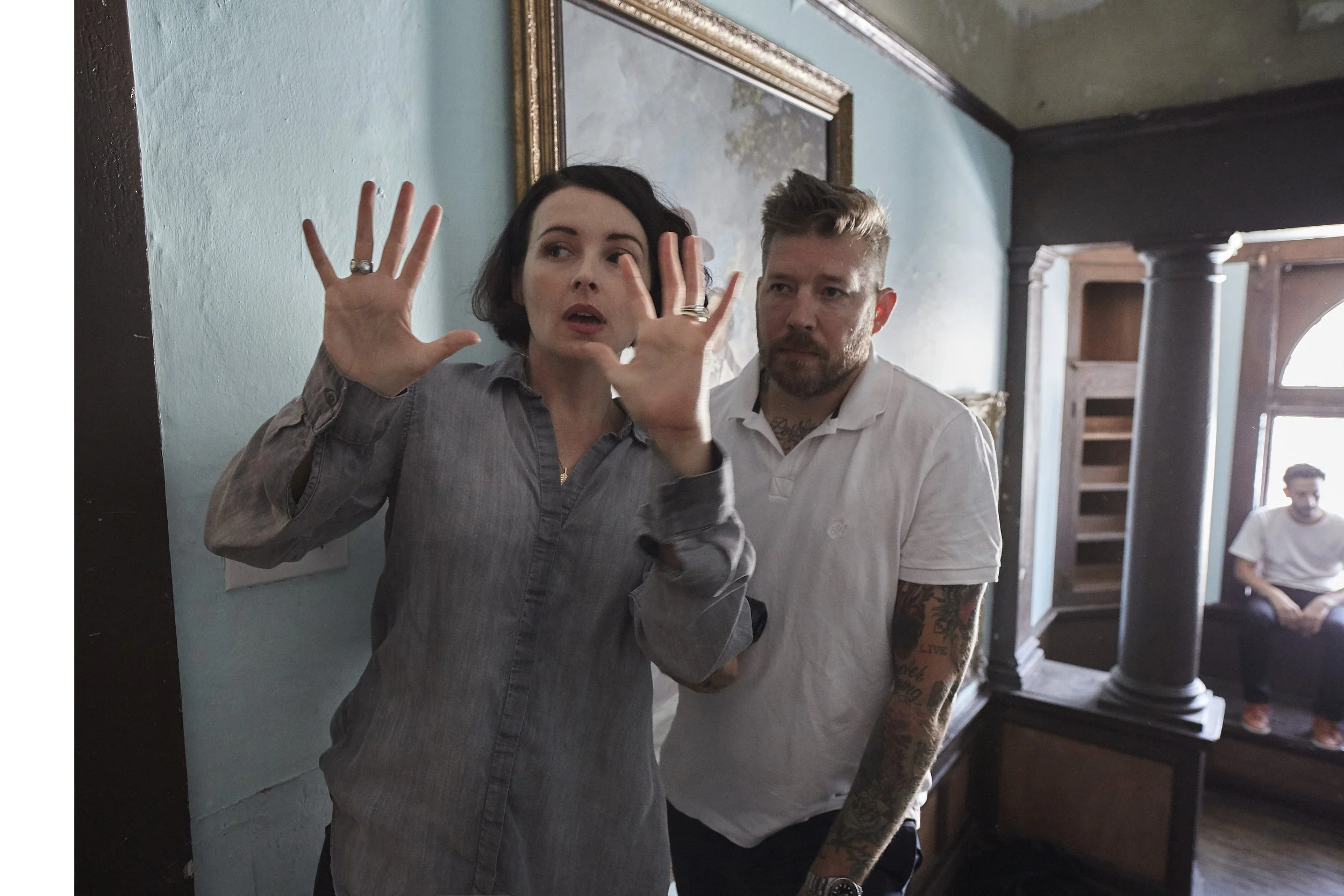A woman with dark hair and tattoos on her right arm is speaking and gesturing with her hands, while a man with light hair and tattoos on his neck looks on. They are inside a room with a blue wall, a gold-framed mirror, and wooden shelves, and appear to be having a conversation or discussion.