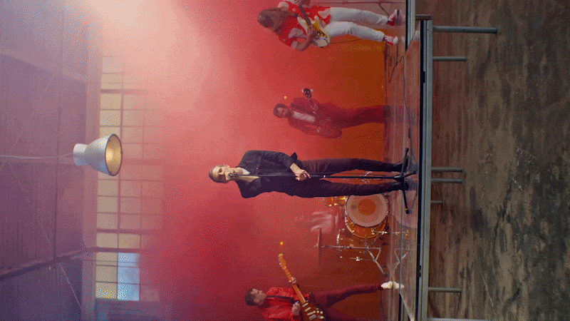 A man in a black suit speaking into a microphone on a stage with a drum set and musicians in the background, surrounded by red fog.