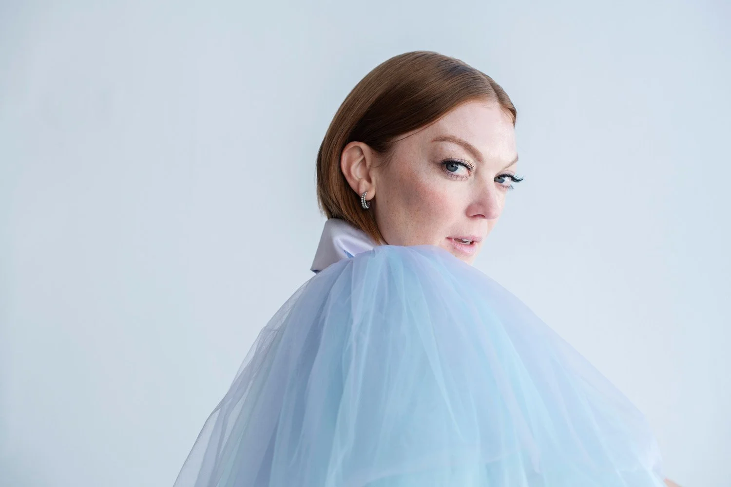 A woman with short red hair, blue eyes, and earrings looking over her shoulder at the camera, wearing a white shirt and a light blue tulle fabric.