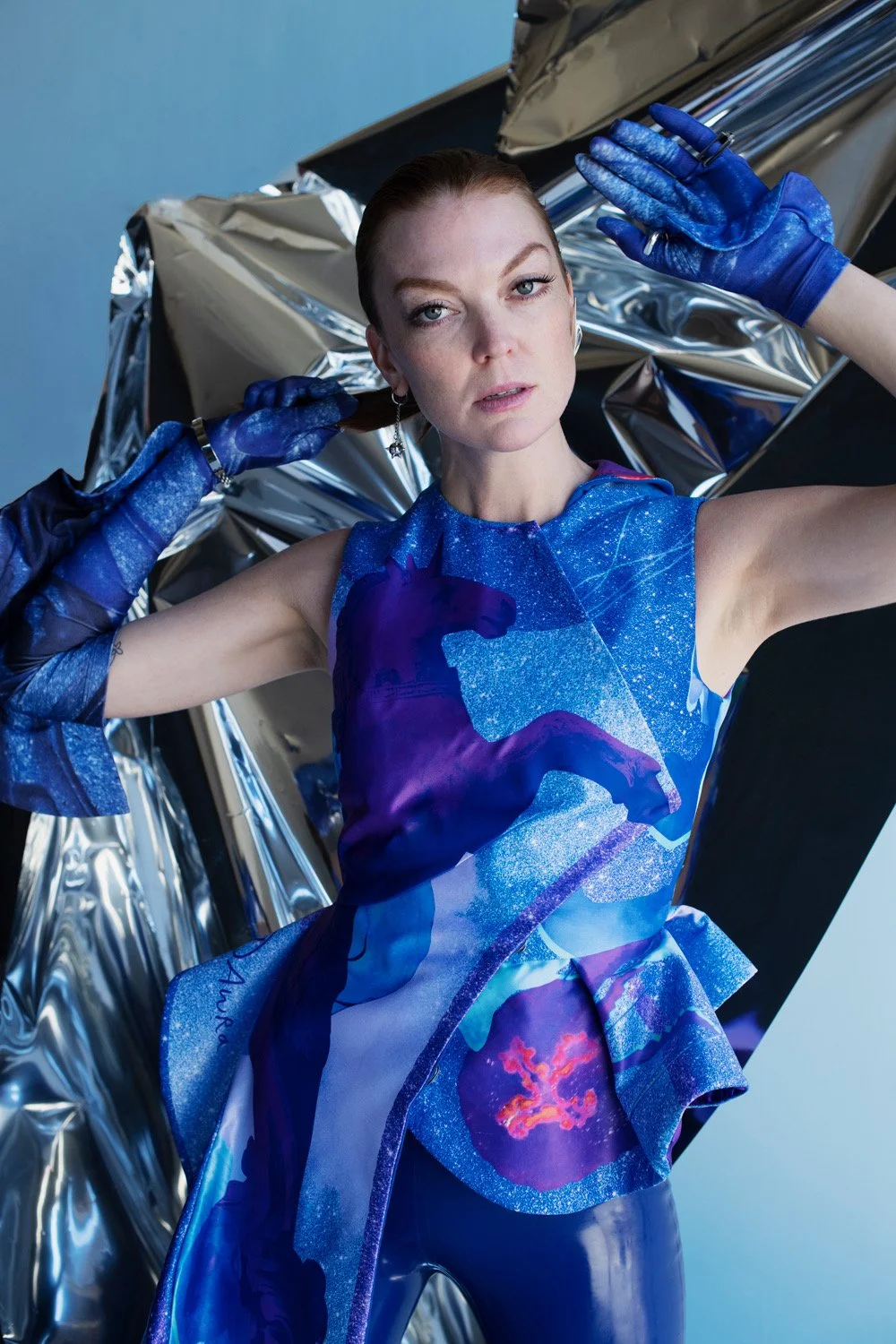 A woman wearing a futuristic, space-themed blue dress with a horse silhouette and galaxy design, along with matching blue gloves, is posing in front of a metallic backdrop.