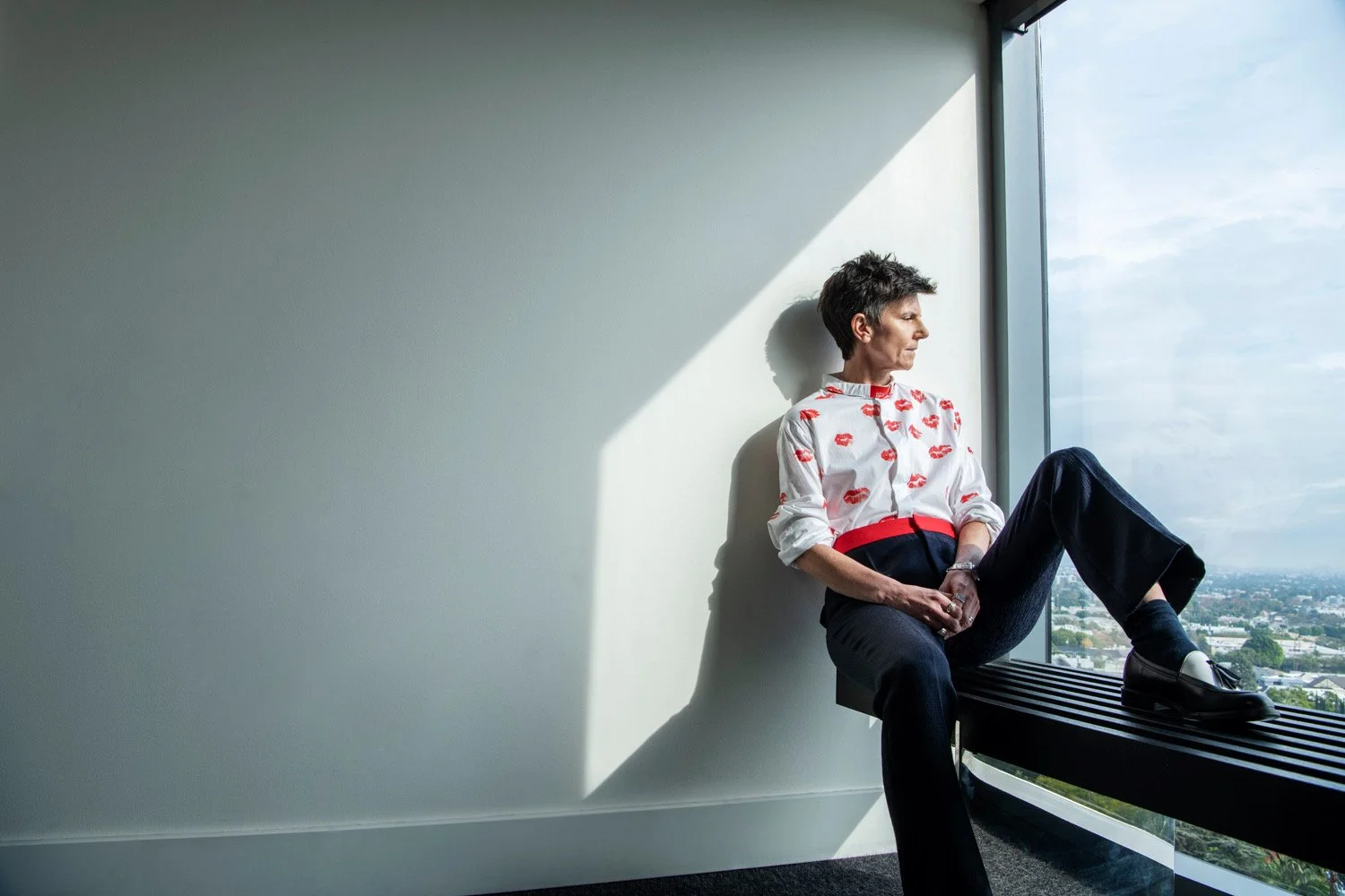 A person with short dark hair sitting on a window seat in a modern office, looking out at the city skyline on a bright day. They are wearing a white shirt with red lipstick kiss prints and dark pants.