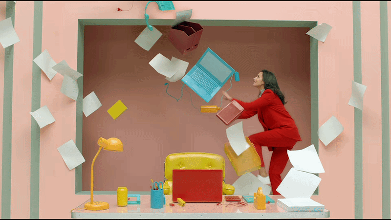 A woman in a red outfit appears to be falling into or through a colorful, whimsical office or studio space, with papers and office supplies flying around and a yellow chair and desk in the foreground.