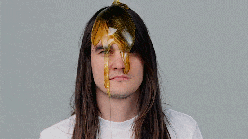 A young man with long brown hair, wearing a white shirt, has honey dripping from a spoon held above his face.