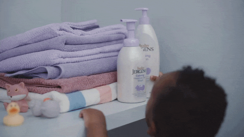 A baby reaching for bottles of Johnson's baby lotion and wash on a changing table, with folded towels and plush toys nearby.