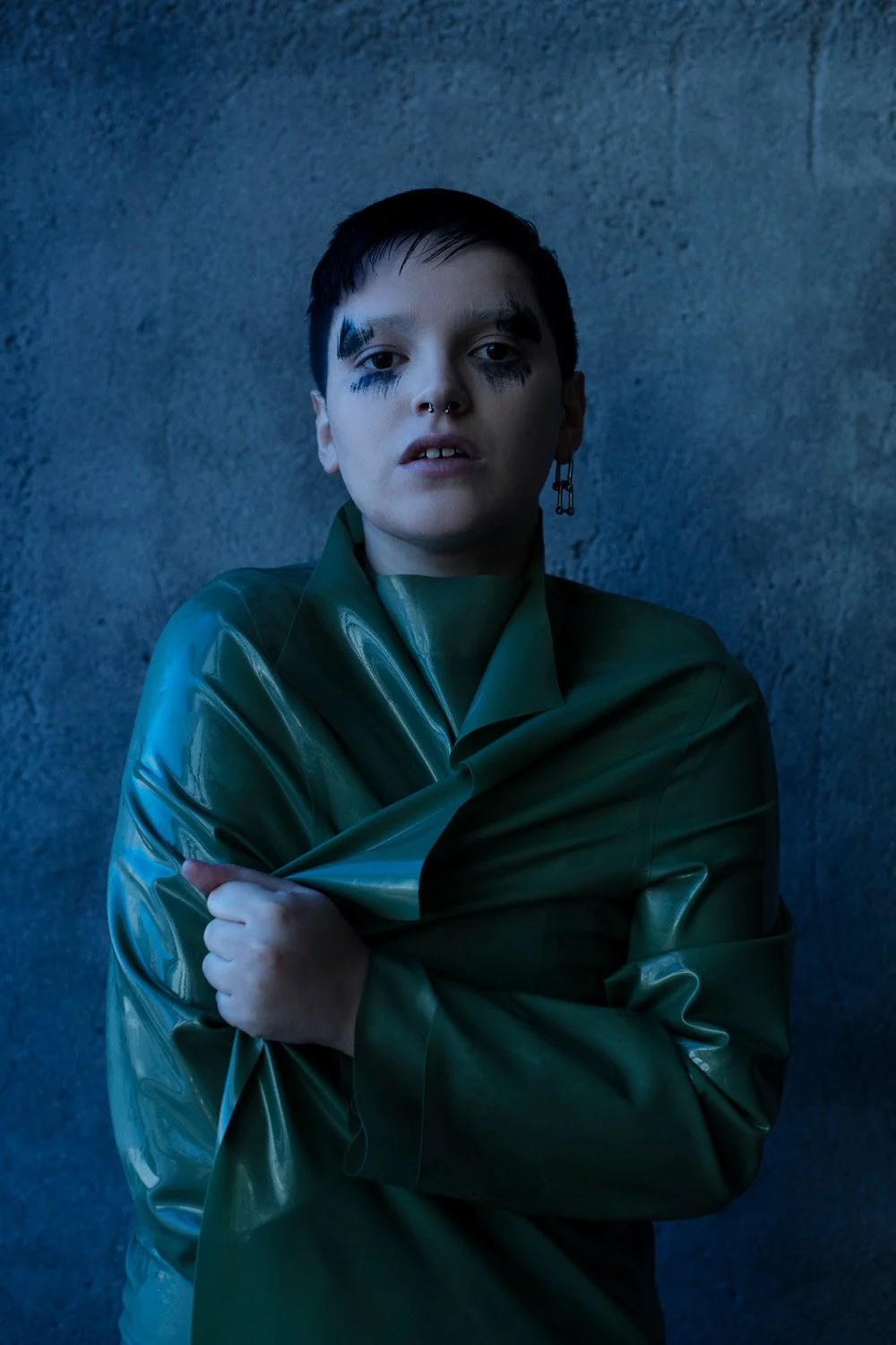 Young person with makeup and an earring, wearing a green raincoat, standing against a gray concrete wall.
