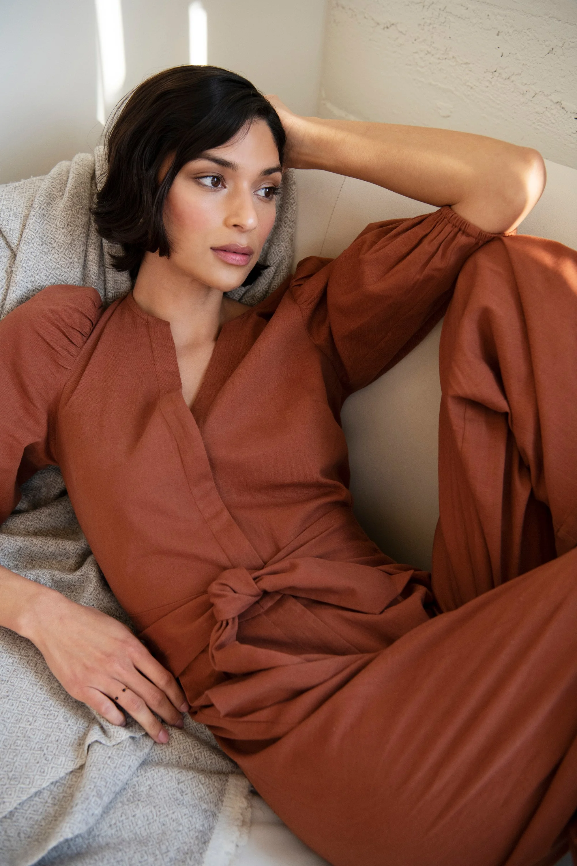 A woman with short dark hair and light makeup, wearing a rust-colored dress, lying on a beige couch with textured fabric. She has her hand resting on her head and appears to be deep in thought, looking off to the side, in a softly lit room.