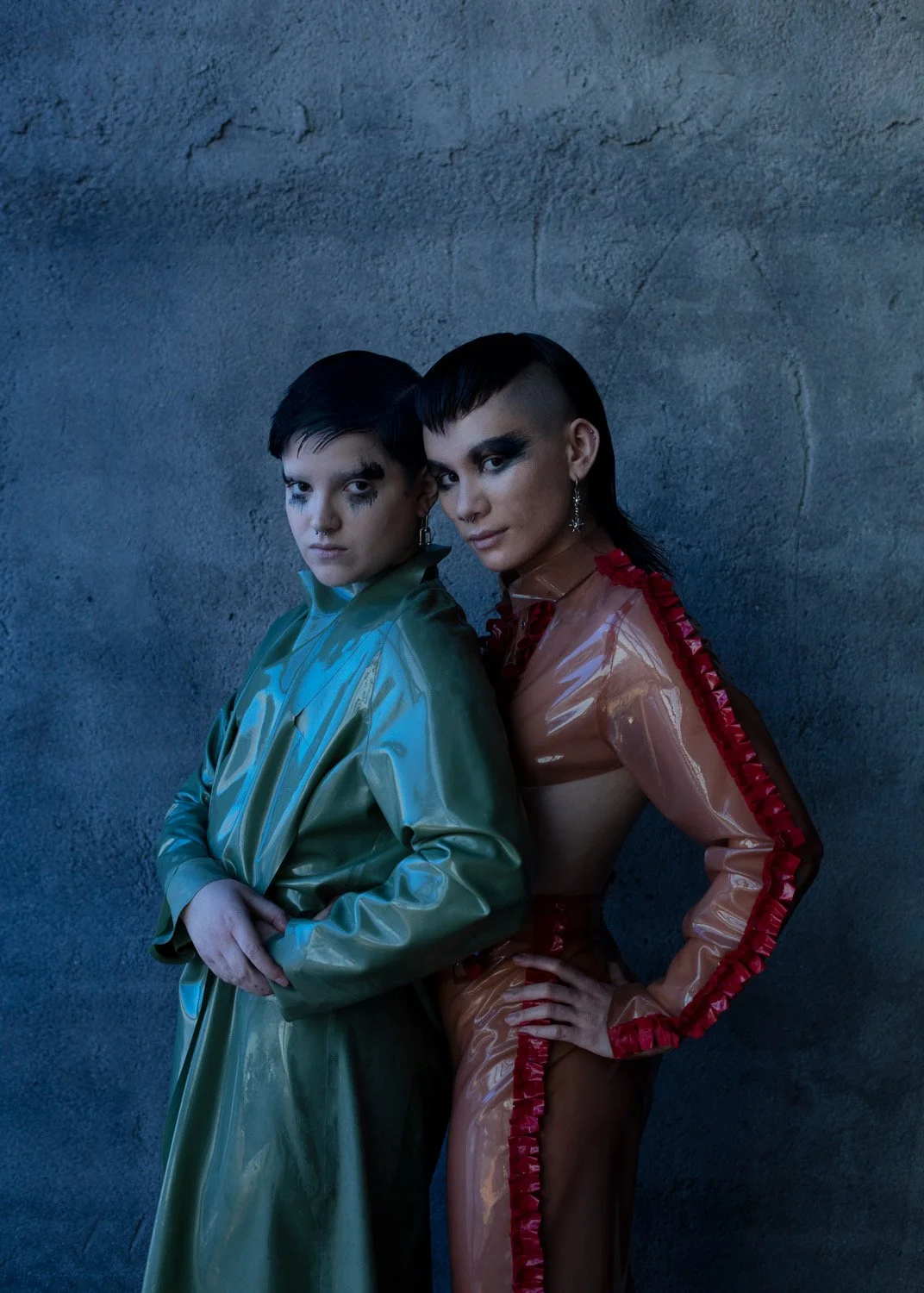 Two women with short black hair and bold makeup posing against a textured gray wall. One wears a shiny green coat, and the other wears a sheer, shiny, peach-colored outfit with red trim.