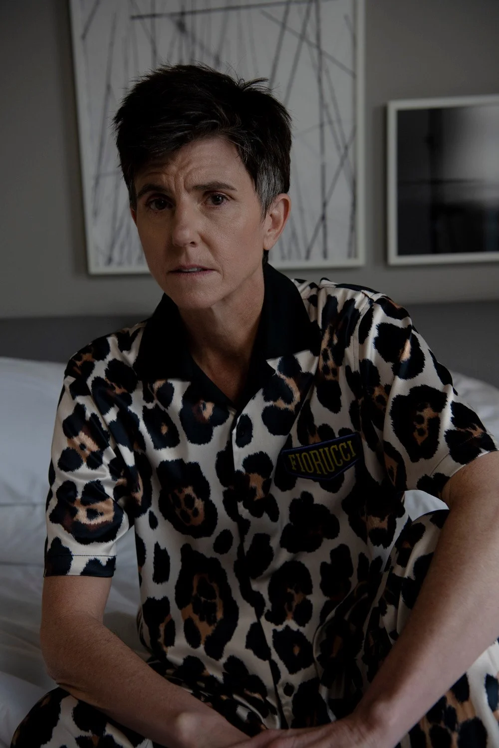 A woman with short dark hair, wearing a leopard print shirt, sitting on a bed in a modern room.