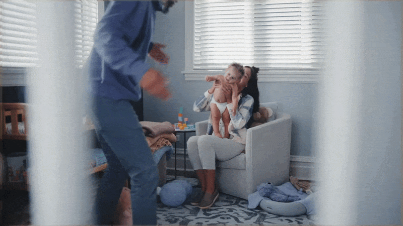 A woman sitting in an armchair holding a smiling toddler, with a man standing nearby in a room with windows and blinds.