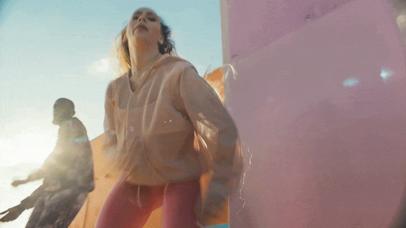 A woman in a peach-colored jacket leans against a pink wall with her head tilted back while a man in a white shirt sits nearby on a beach with orange cliffs in the background.