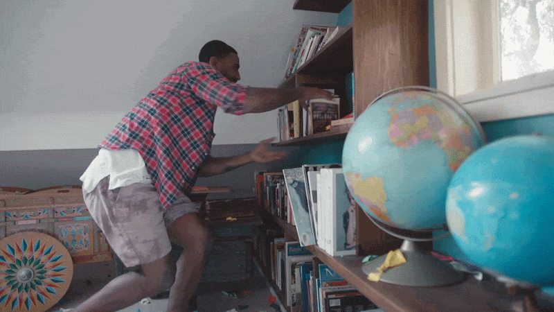Young man in a plaid shirt and shorts organizing books on a bookshelf in a room with a window, globes, and clutter.