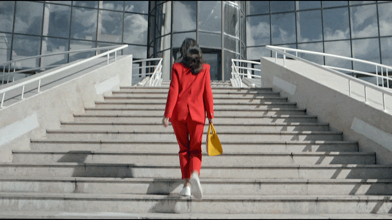 A woman in a red suit with white shoes carrying a yellow handbag walks up a staircase outside of a modern glass building.