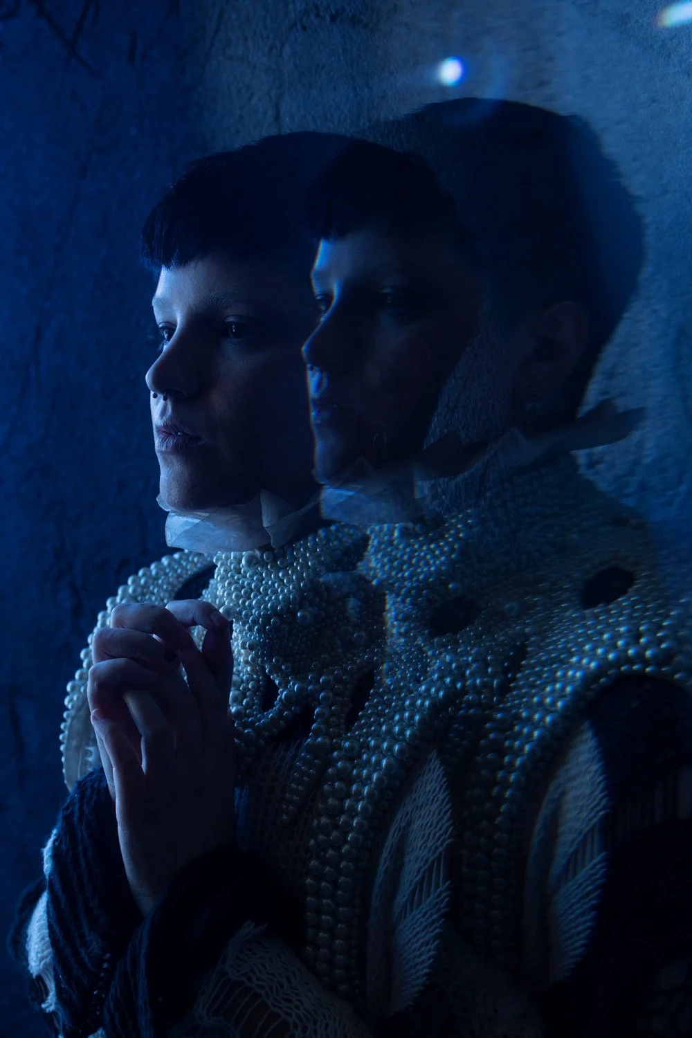 A person with short dark hair and pale skin, wearing a pearl or bead jewelry and a textured garment, standing with hands clasped and looking thoughtfully to the side in front of a textured dark wall, reflected in a mirror.