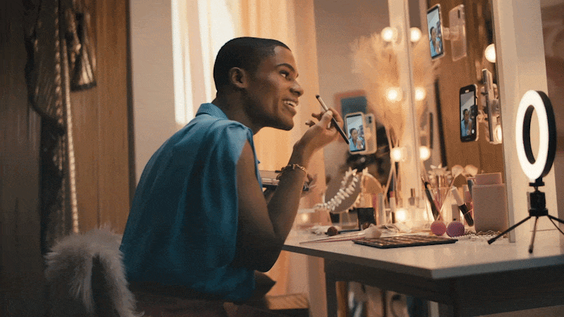 Young woman doing makeup while taking selfies in a well-lit vanity mirror, with a ring light, makeup products, and phone mounted on the mirror.