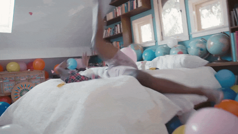 Child jumping on a bed surrounded by colorful balloons in a cozy bedroom with bookshelves and windows.