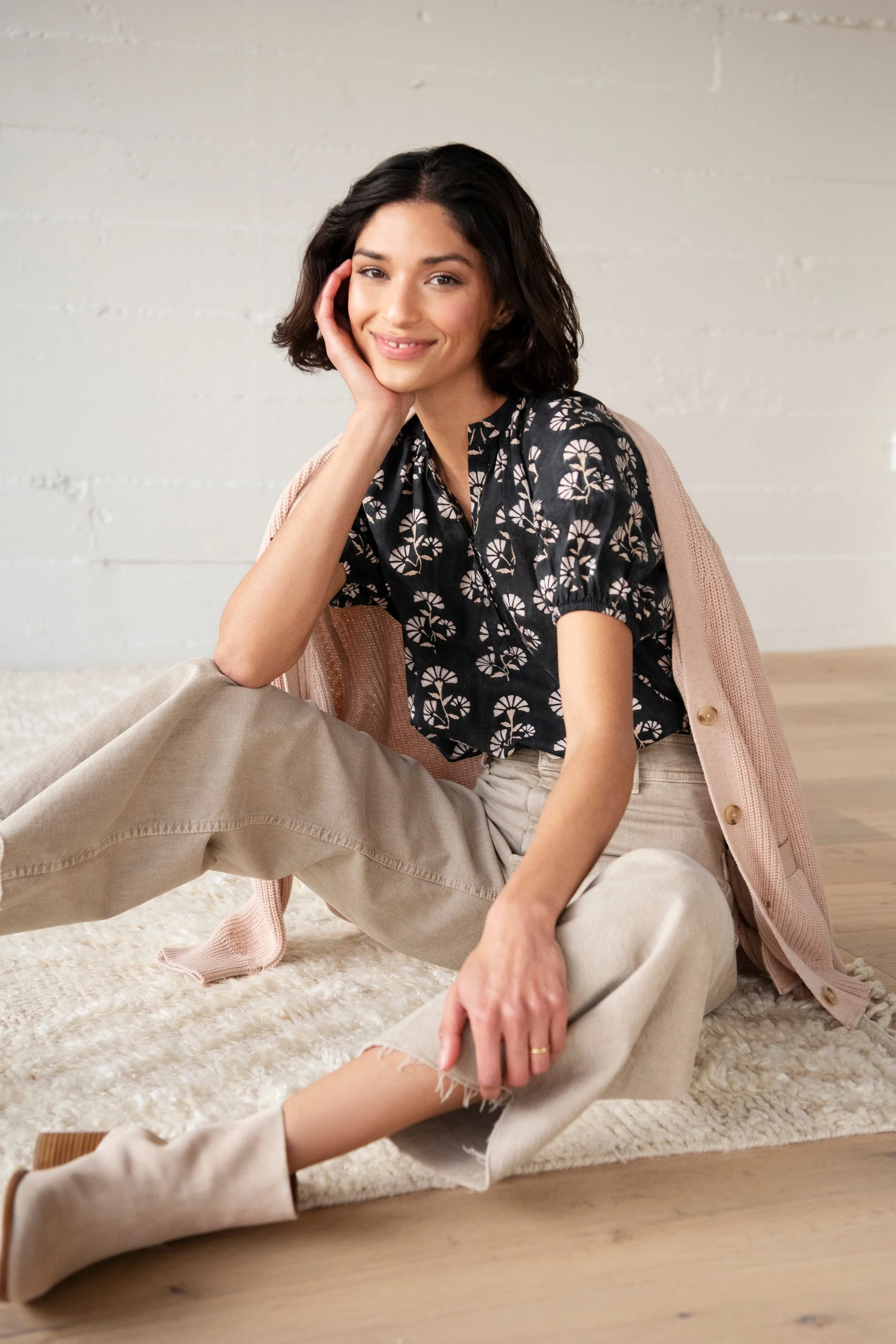 A young woman with dark hair sitting on the floor on a cream-colored rug, smiling at the camera, with one hand resting on her face and the other on her knee, dressed in beige pants and a patterned black top, with a pink cardigan draped over her shoul