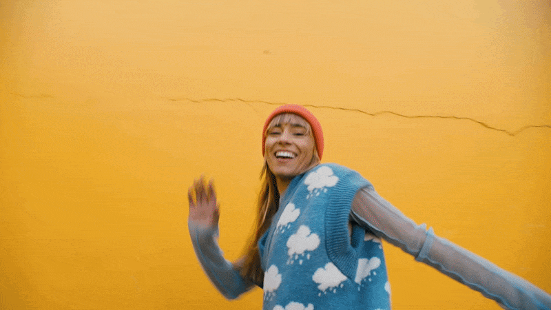 A young woman with a bright smile, wearing a light blue sweater with white clouds, an orange beanie, and a sheer shirt underneath, standing in front of a yellow wall.