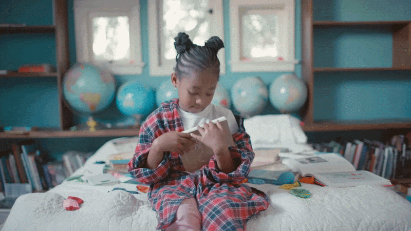 A young girl sitting on a bed in a bedroom, wearing pajamas, focused on a tablet device she is holding in her hands.