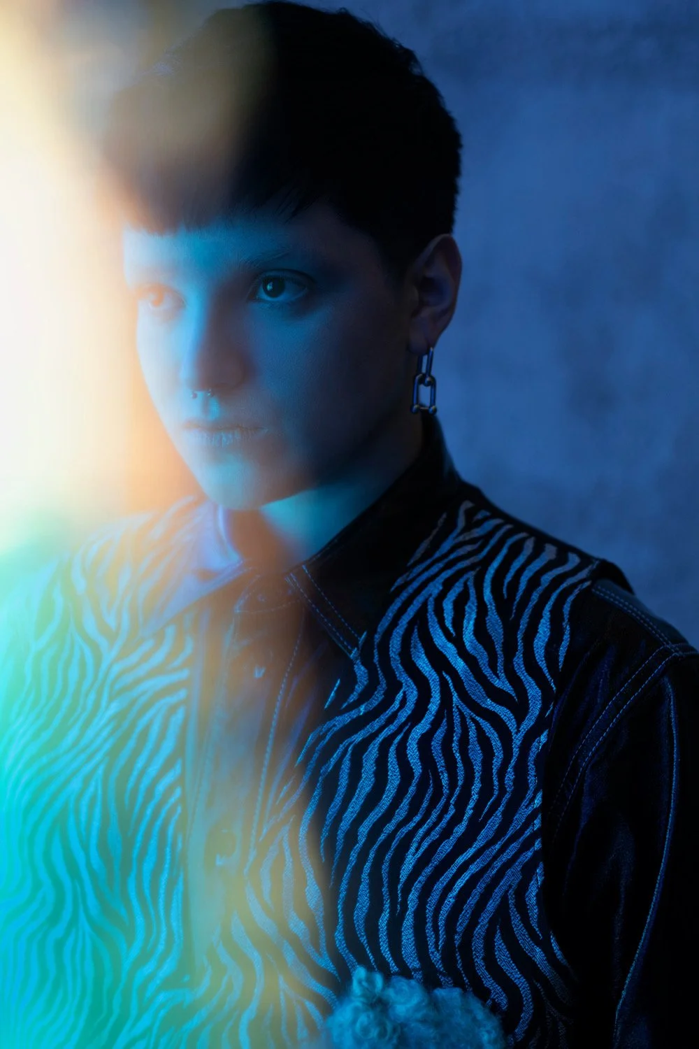 A young person with short dark hair, wearing a black and white zebra-patterned jacket and a dangling earring, gazing thoughtfully into the distance with a blue-toned background and colorful light effects.