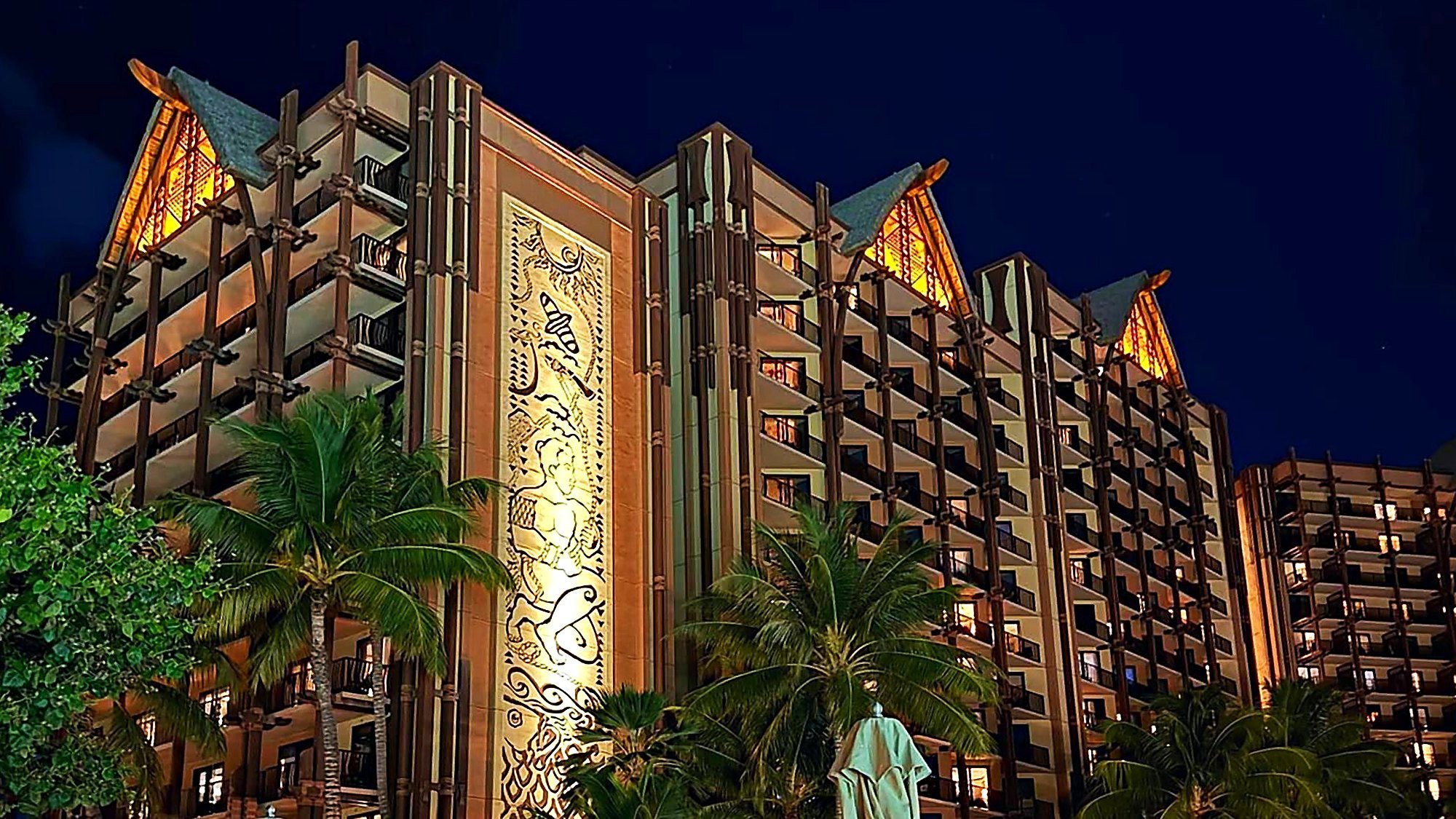 Aulani resort tower at night with glowing gables, a carved mural and palm trees