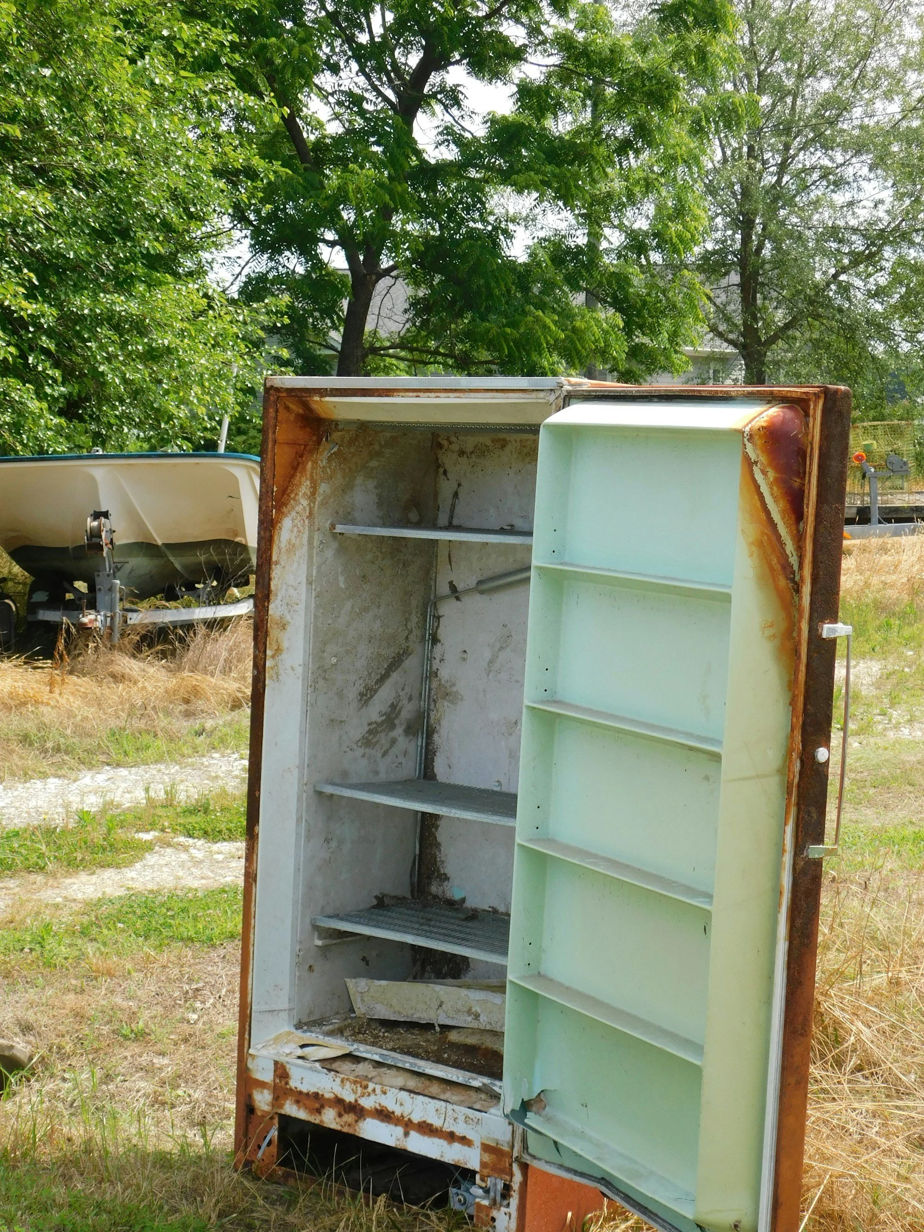 An old, rusty refrigerator stands outdoors with its door open, revealing an empty interior, pending haul away.