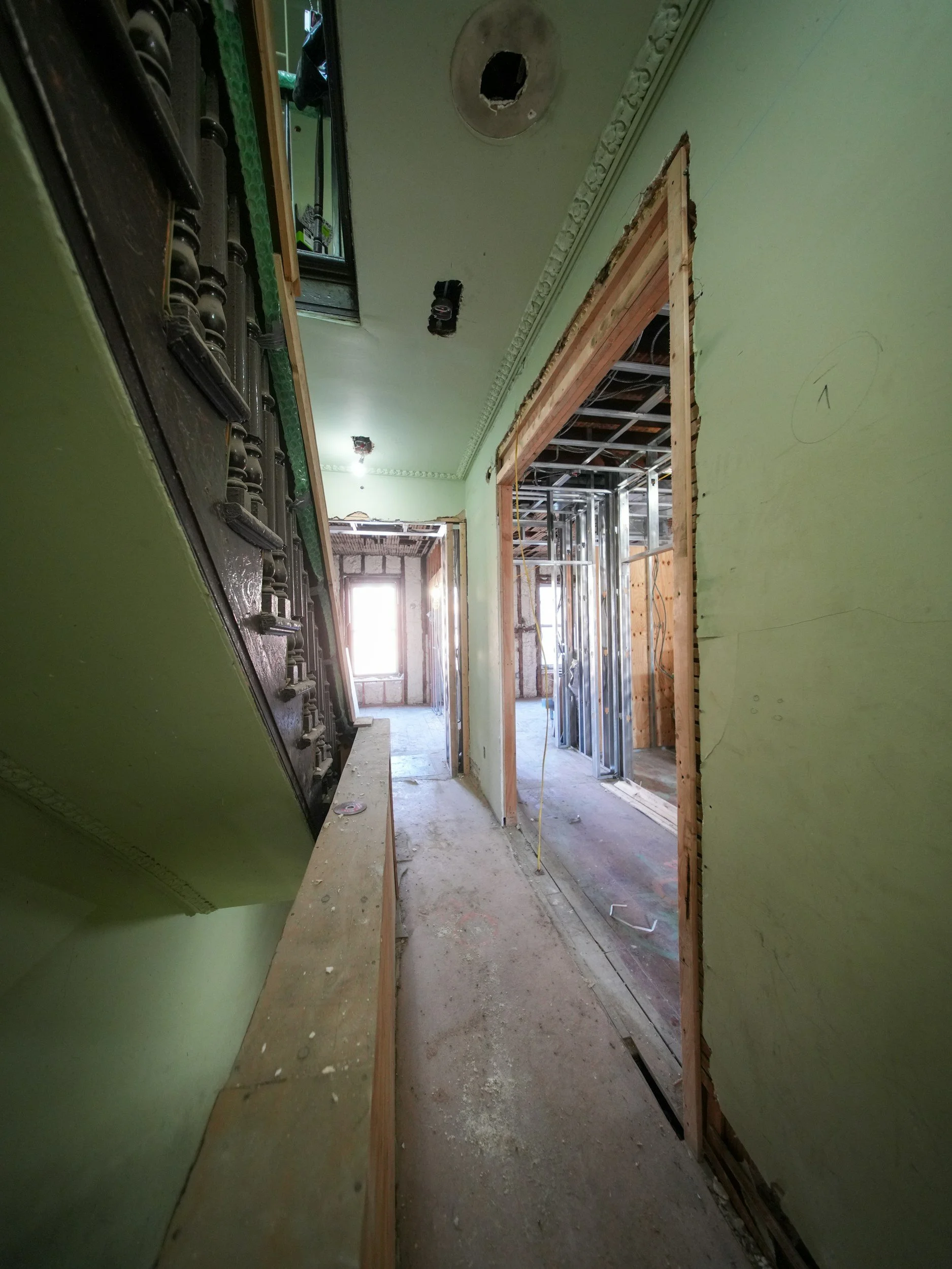 Interior of a building under renovation showing green painted walls, exposed framing around doorways, a partially removed ceiling, and construction materials on the floor.