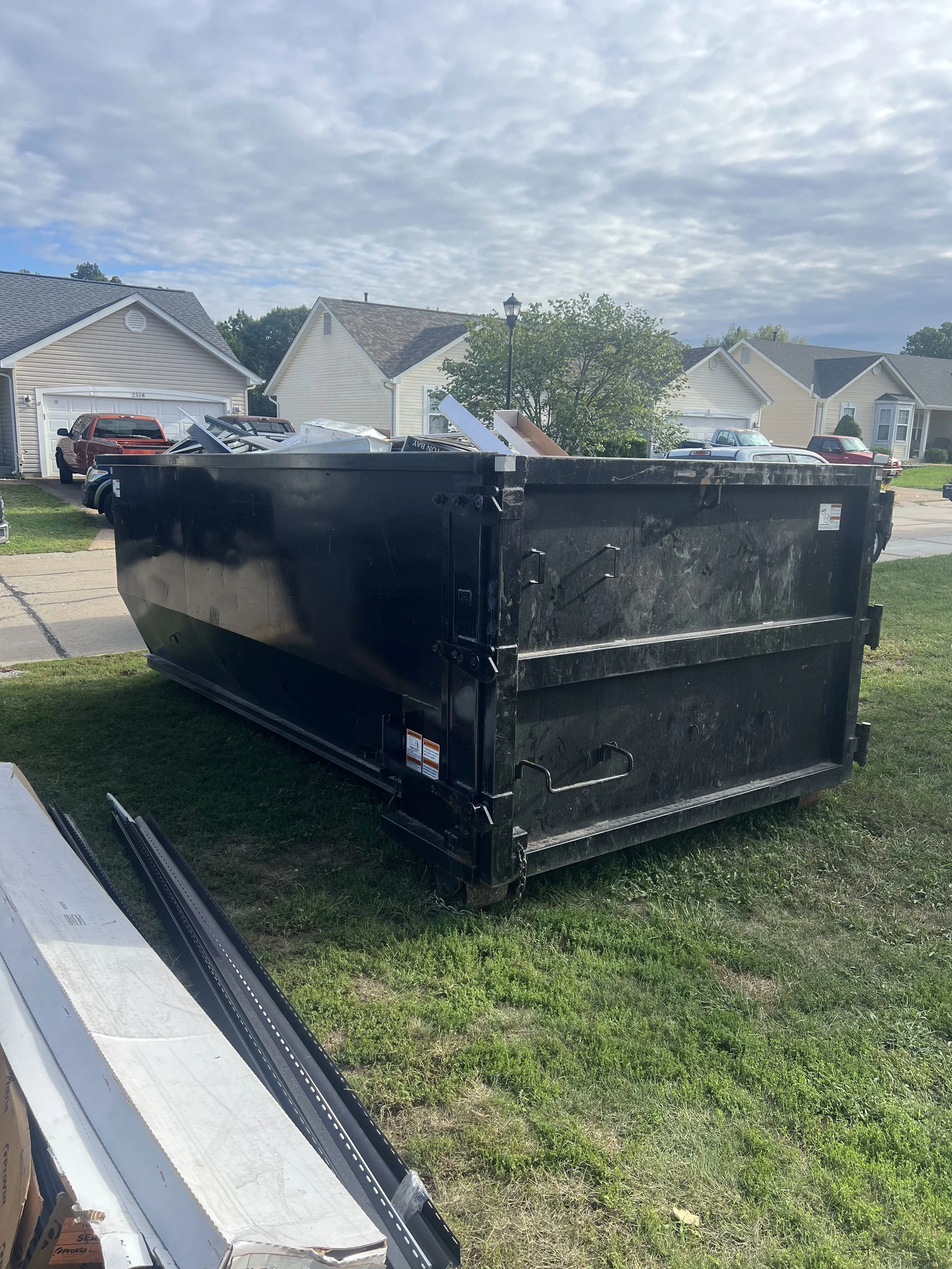 A black dumpster filled with construction debris on a lawn in a residential neighborhood.