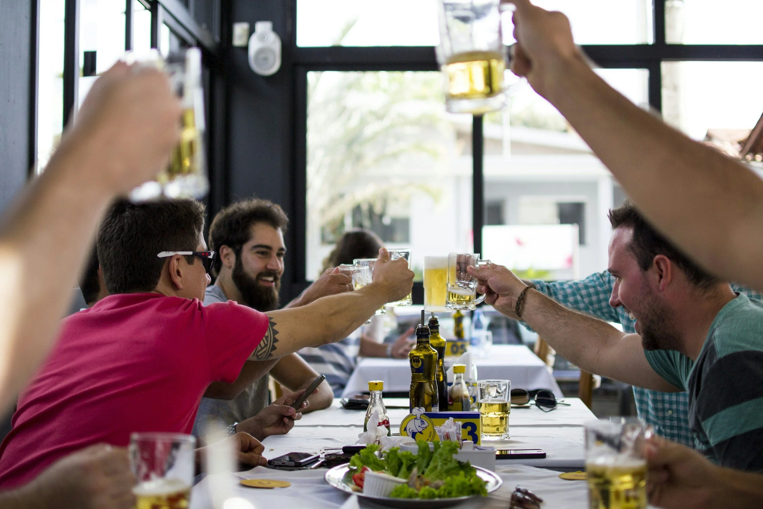 People raising glasses of beer in a toast at a restaurant or cafe with food and condiments on the table.