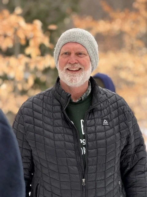 A smiling middle-aged man with a gray beard and mustache wearing a gray knit beanie and a black quilted jacket, standing outdoors with autumn-colored trees in the background.