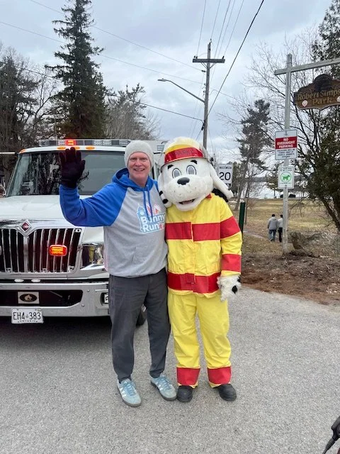 A person wearing a blue hoodie and gray pants standing next to a person in a mascot costume resembling a firefighter dog, both waving, with a fire truck behind them on a street.