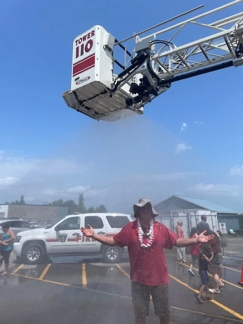 Person standing behind a fire truck with their arms outstretched, smoke rising from the vehicle, and a fire department vehicle in the background on a parking lot.