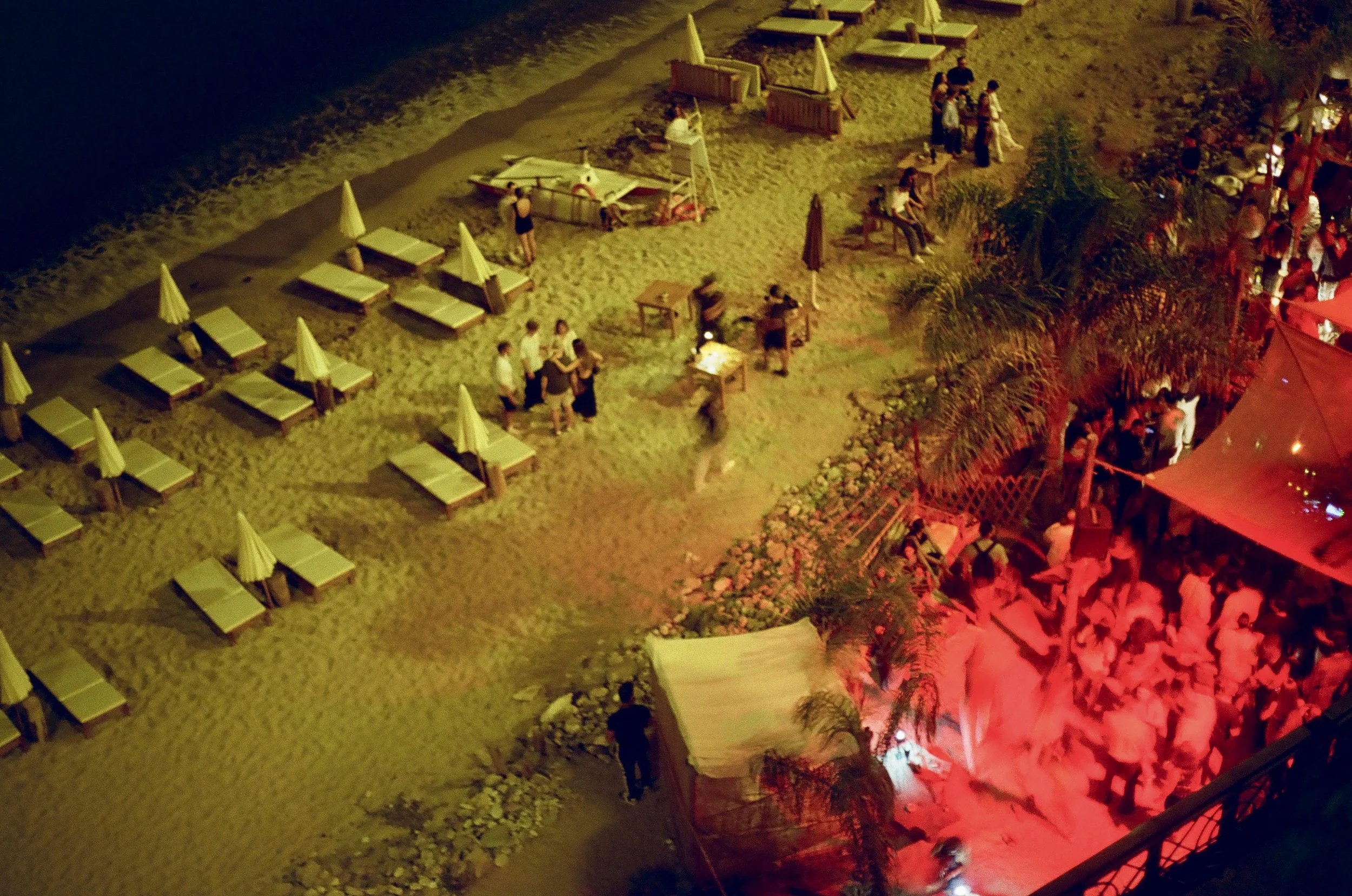 Nighttime scene of a beach area with empty lounge chairs and umbrellas, groups of people standing and sitting, and a red-lit structure with plants nearby.
