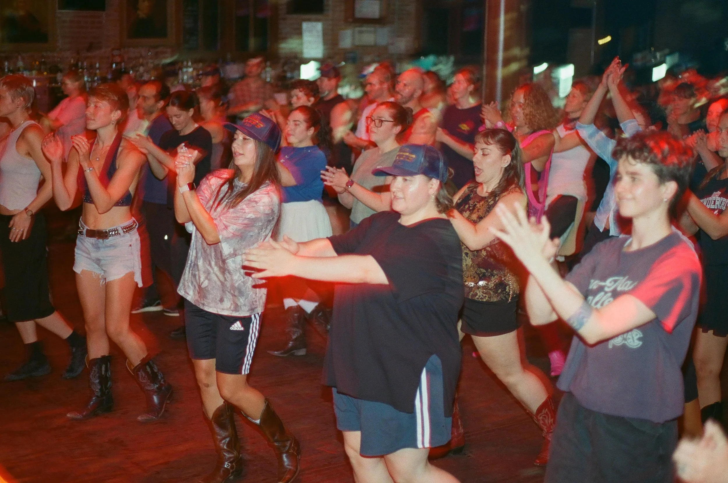A group of people dancing closely together in a lively, crowded indoor venue with red lighting.