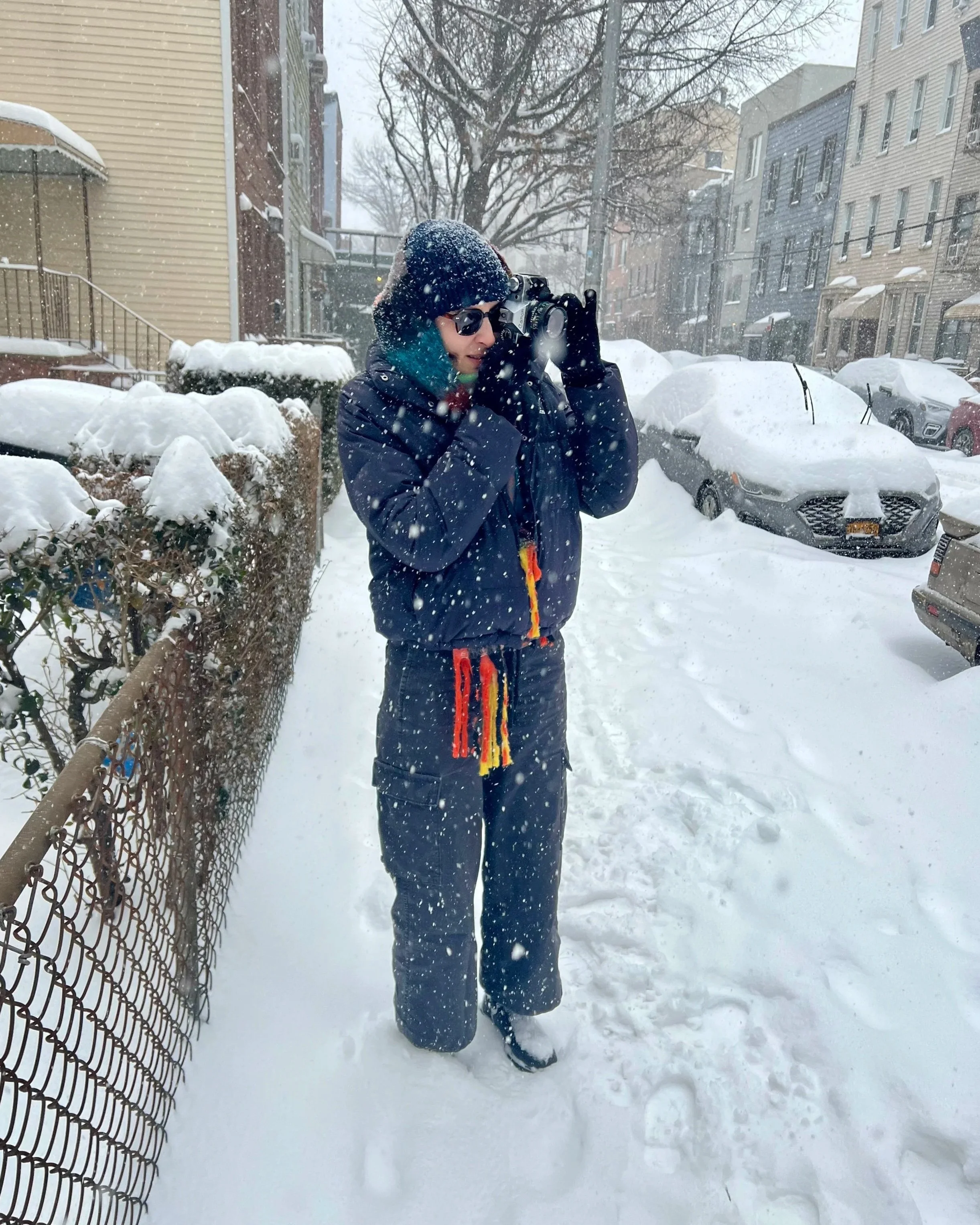 A person standing outdoors in a snowy environment, wearing a dark winter jacket, gloves, sunglasses, and a knit hat with snow on it. They are holding a camera up to their face, taking a photo. Snow covers the parked cars and the ground, and snow is falling. Residential buildings and a leafless tree are visible in the background.