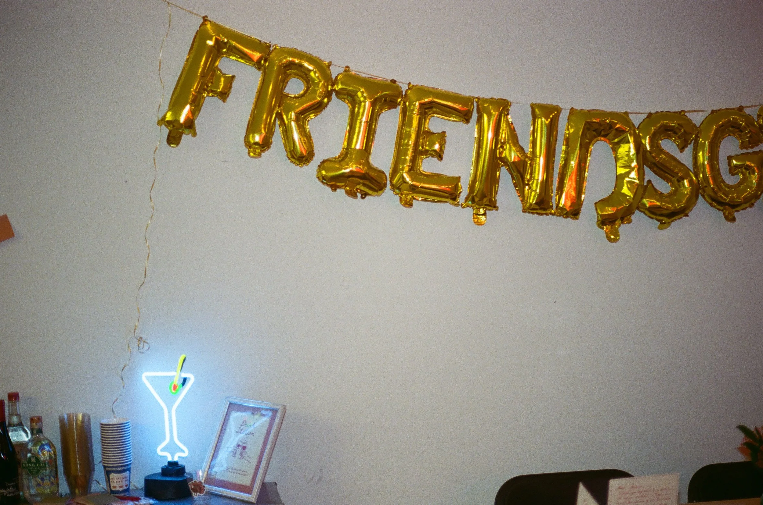 Golden balloon banner spelling 'FRIENDSHIP' hanging on a wall in a party setting, with a bar area at the bottom featuring bottles, cups, and a neon drink sign.