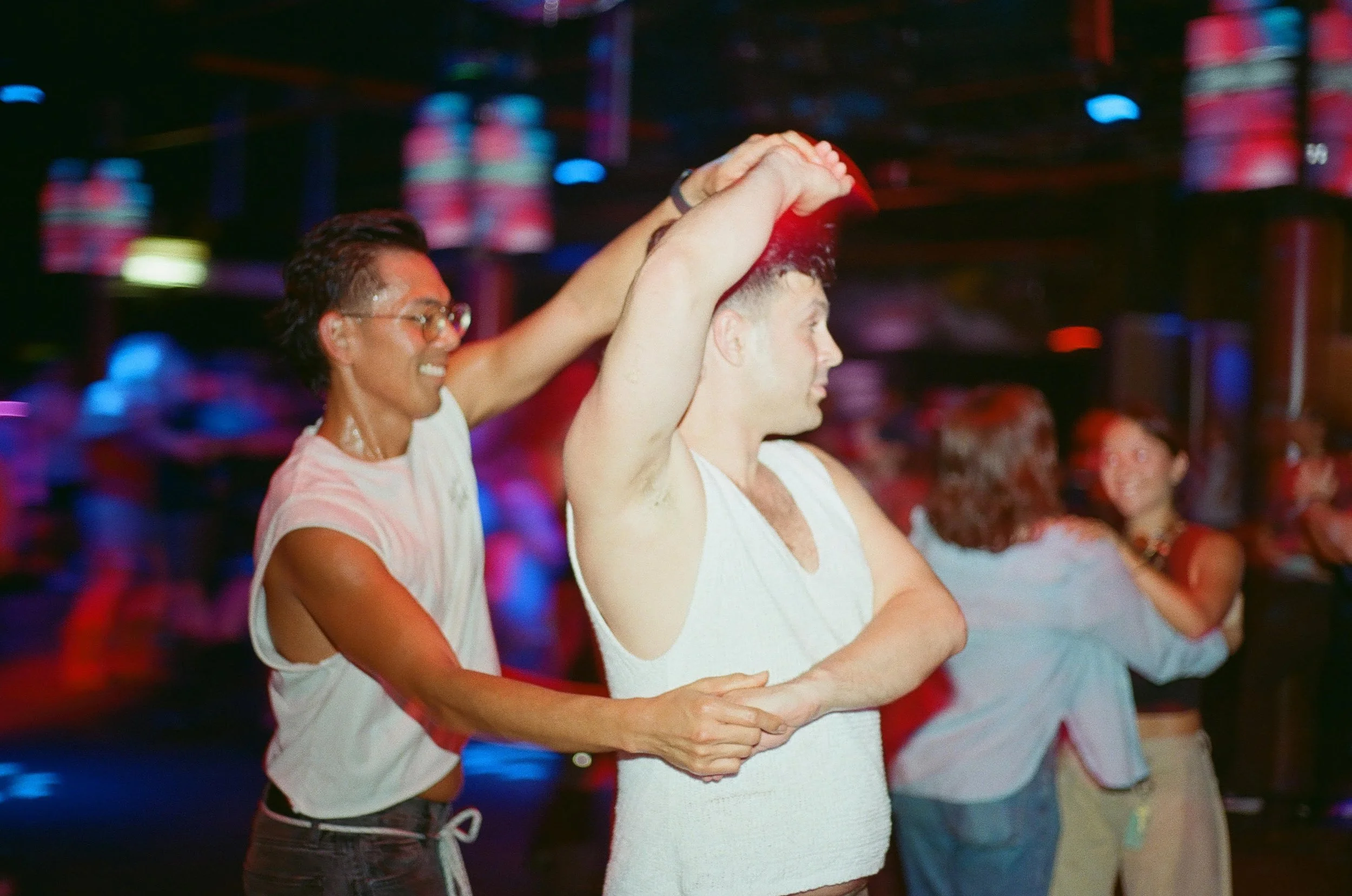 A group of people dancing in a line, with a man and woman in the foreground holding each other's hands and smiling, in a lively setting with colorful lights.