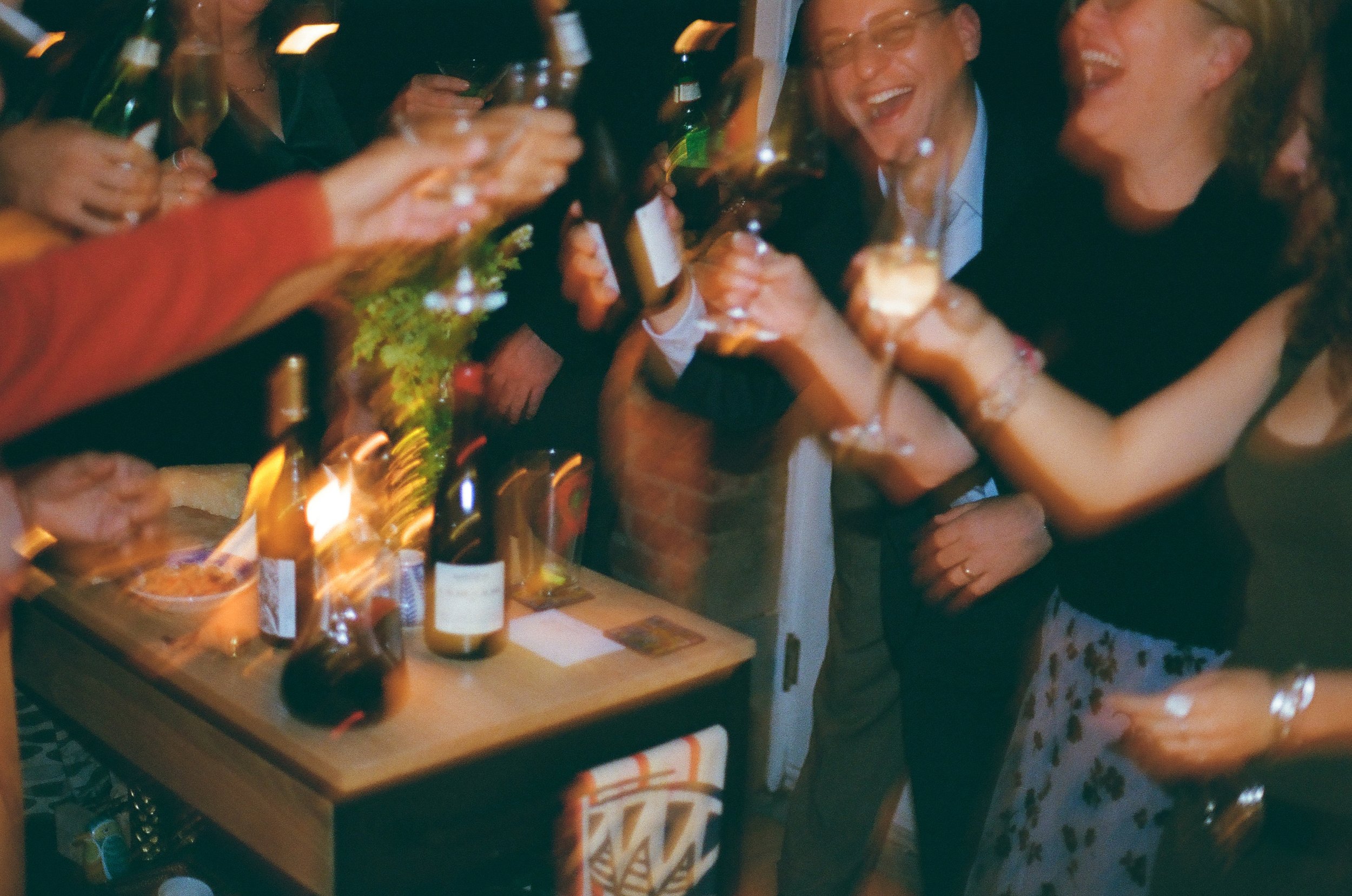 People celebrating at a party, raising glasses, with a table of drinks and snacks, smiling and laughing.