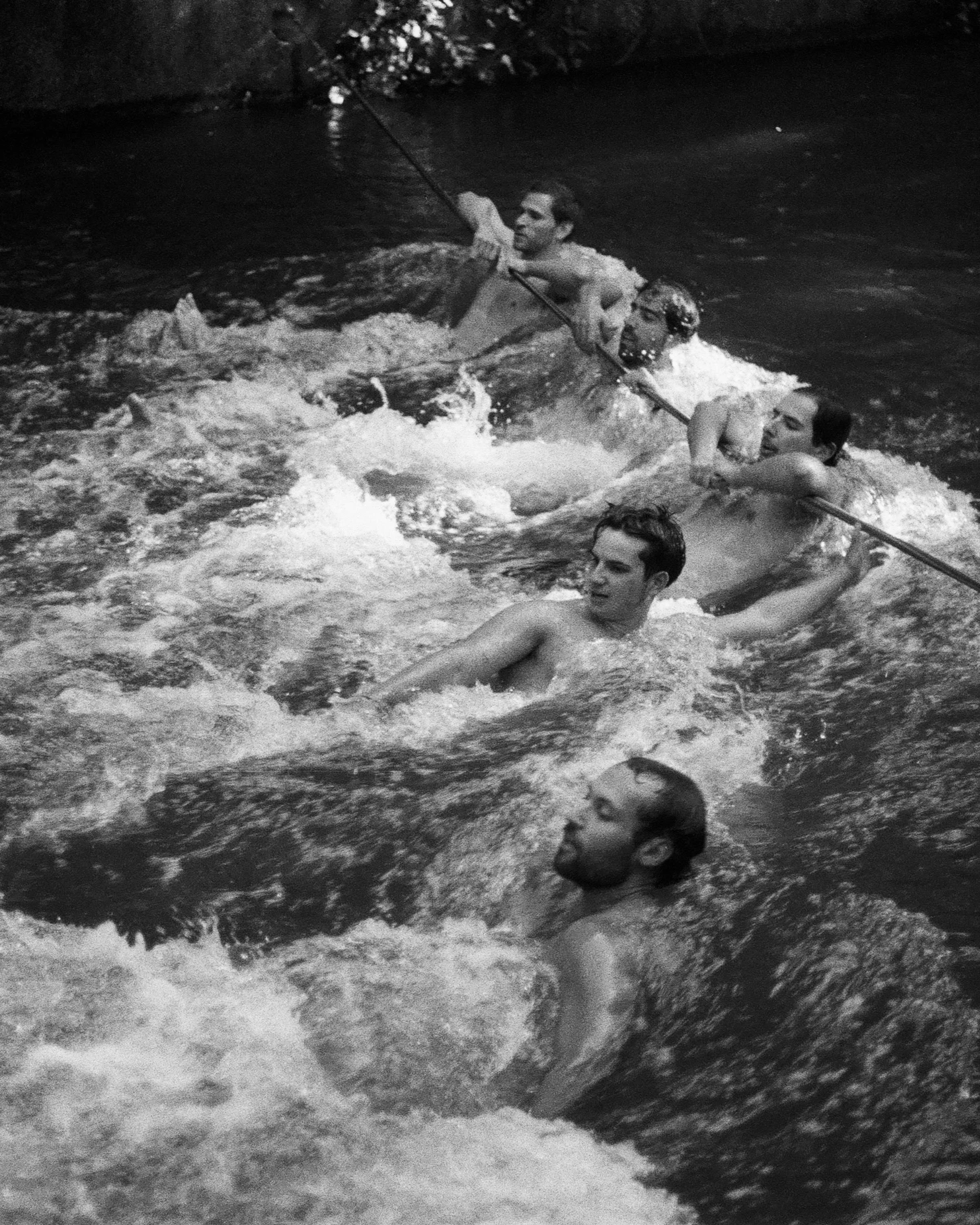 Four men are swimming in a river, holding on to a rope, with one man using a fishing rod. The image is in black and white.