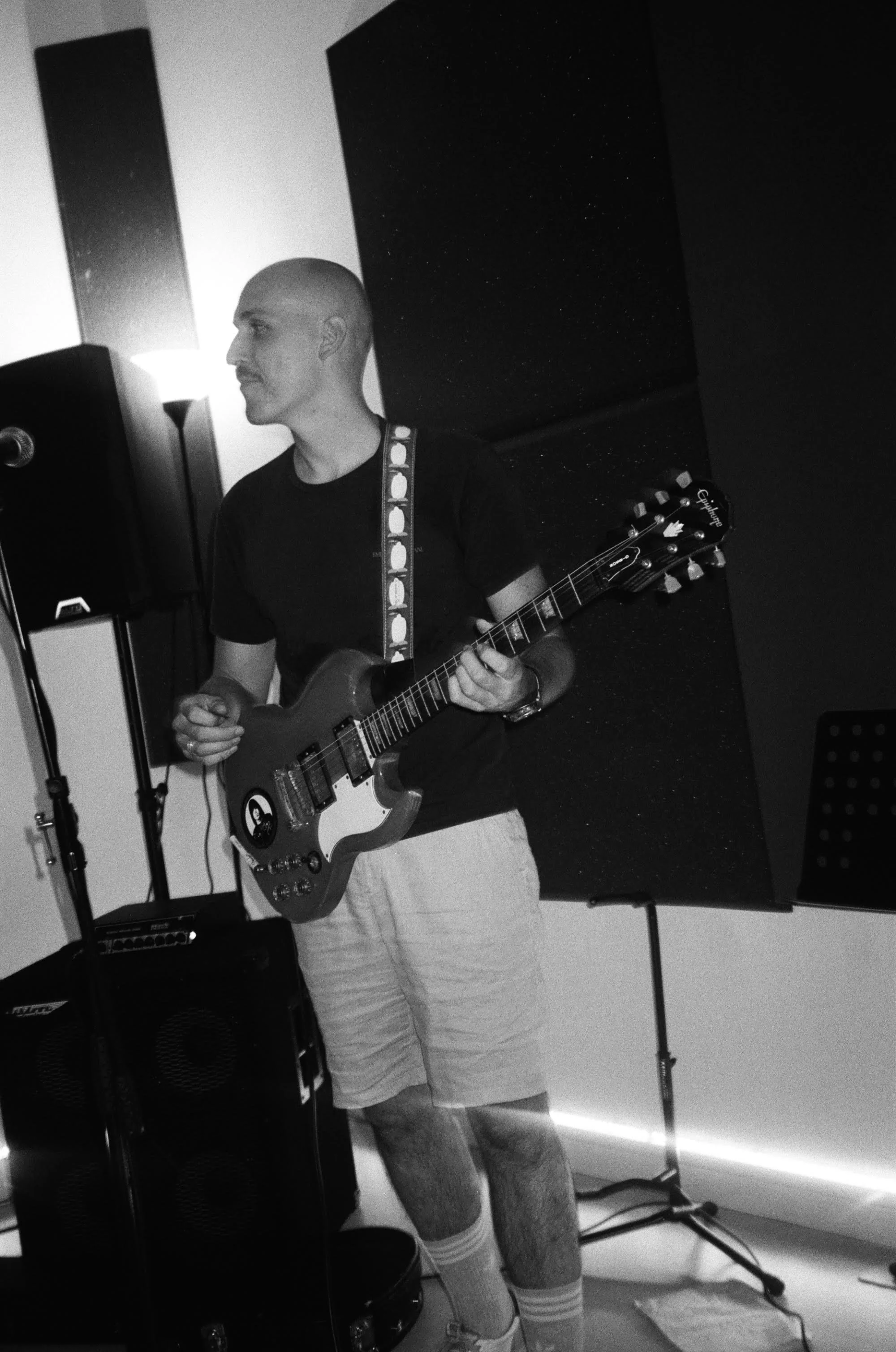 A man with a shaved head playing an electric guitar in a music studio, wearing a black t-shirt, shorts, and striped socks.